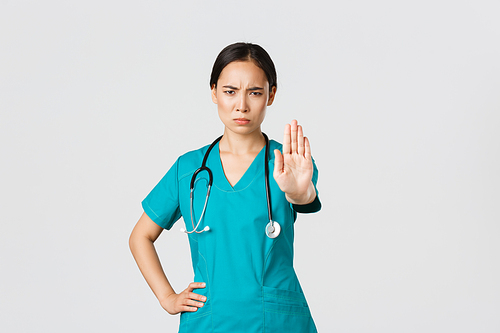 Covid-19, healthcare workers, pandemic concept. Angry serious-looking asian doctor, female physician or nurse in scrubs frowning displeased, extend hand to show stop, disagree, prohibit or forbid.
