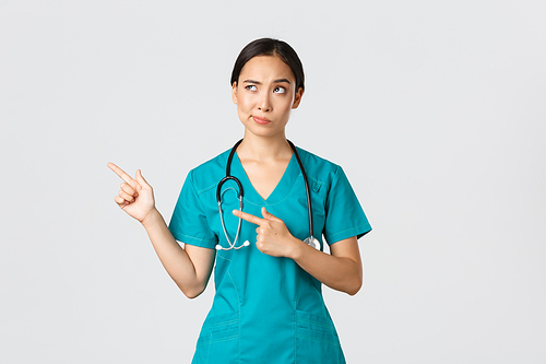 Covid-19, healthcare workers, pandemic concept. Skeptical and doubtful pretty asian doctor, nurse in scrubs smirk, pointing upper left corner, looking with reluctant hesitant face, white background.