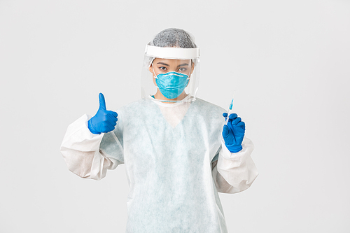 Covid-19, coronavirus disease, healthcare workers concept. Confident serious female asian doctor in personal protective equipment, show thumb-up and hold syringe with vaccine, white background.