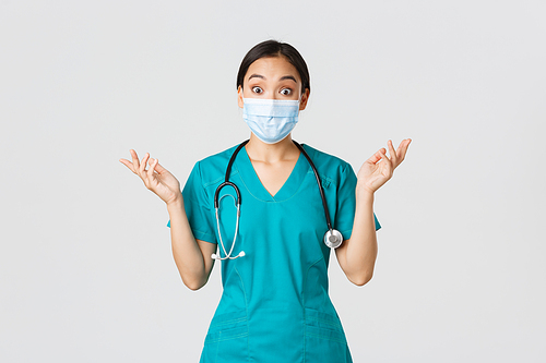 Covid-19, coronavirus disease, healthcare workers concept. Surprised and amazed asian female nurse, physician in medical mask and scrubs raising hands up excited, hear amazing news, white background.