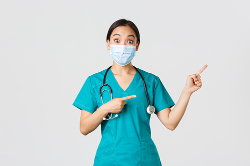 Covid-19, coronavirus disease, healthcare workers concept. Enthusiastic happy asian female doctor, physician or nurse in medical mask and scrubs, pointing upper right corner, show awesome promo.