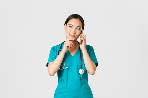 Covid-19, healthcare workers and preventing virus concept. Portrait of smiling asian female doctor, intern in scrubs talking on phone and looking thoughtful, thinking or making choice.