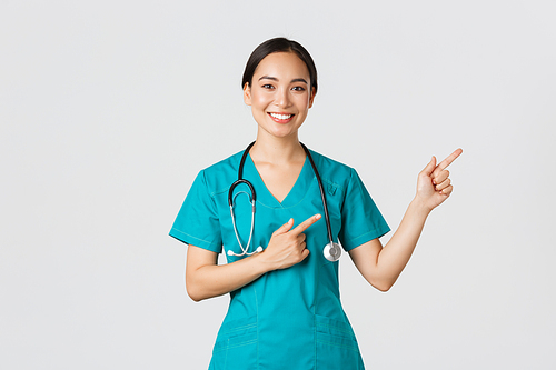 Covid-19, healthcare workers, pandemic concept. Smiling beautiful asian female nurse, intern pointing fingers upper right corner, inviting for checkup, online clinic appointment, white background.