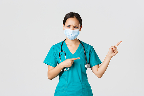 Covid-19, coronavirus disease, healthcare workers concept. Frowning suspicious or frustrated asian female doctor, wear medical mask and scrubs, pointing upper right corner, white background.