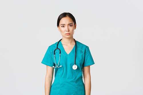 Covid-19, healthcare workers, pandemic concept. Exhausted young asian female nurse, doctor looking tired after shift in hospital, looking sad with fatigue, standing white background in scrubs.