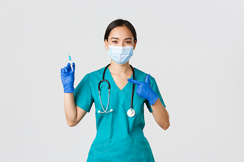 Covid-19, coronavirus disease, healthcare workers concept. Confident friendly-looking asian female doctor, physician in medical mask and scrubs pointing finger at syringe with vaccine.