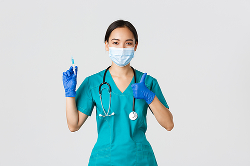 Covid-19, coronavirus disease, healthcare workers concept. Pleasant smiling asian female doctor, physician in medical mask and gloves, show thumb-up, hold syringe with vaccine.