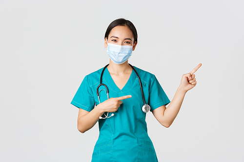 Covid-19, coronavirus disease, healthcare workers concept. Pleasant asian female doctor, medical employee in scrubs and mask, pointing fingers upper right corner, showing advertisement.