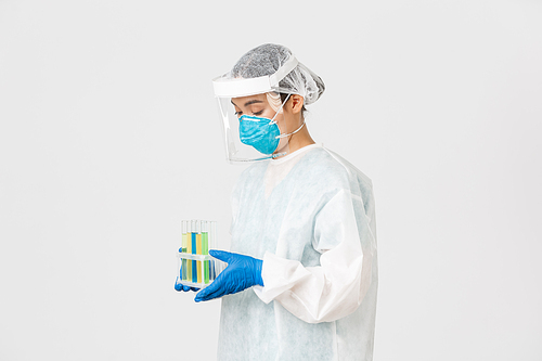 Covid-19, coronavirus disease, healthcare workers concept. Side view of careful serious asian female doctor, tech lab employee in personal protective equipment holding test-tube probes of vaccines.