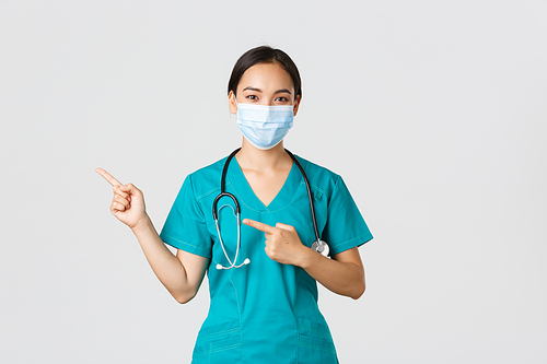 Covid-19, coronavirus disease, healthcare workers concept. Professional confident asian female physician, doctor in medical mask and scrubs pointing fingers left, showing advertisement.