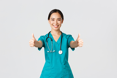 Covid-19, healthcare workers, pandemic concept. Smiling pretty asian female doctor, nurse in scrubs ensure everything good, showing thumbs-up in approval, agree or like something, white background.