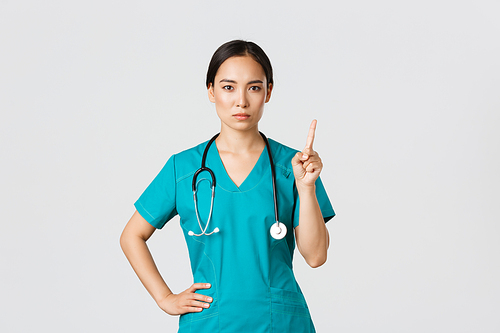 Covid-19, healthcare workers, pandemic concept. Serious-looking professional female asian doctor in scrubs, physician shaking finger in prohibition, warning gesture, forbid something.
