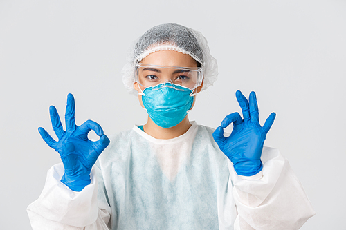 Covid-19, coronavirus disease, healthcare workers concept. Close-up of pleased serious-looking asian female in personal protective equipment show okay gesture, approve or guarantee something.