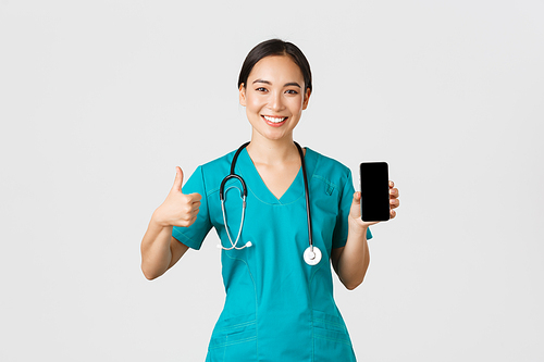 Covid-19, healthcare workers and online medicine concept. Portrait of young asian female physician, doctor in scrubs show thumbs-up and smartphone screen, promote application, white background.