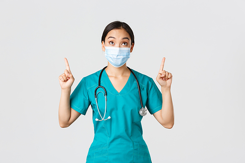 Covid-19, coronavirus disease, healthcare workers concept. Intrigued and excited asian woman doctor, nurse in medical mask and gloves, looking and pointing fingers up, white background.