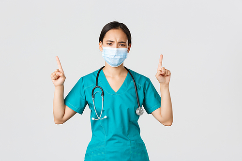Covid-19, coronavirus disease, healthcare workers concept. Gloomy disappointed asian nurse, physician in medical mask and scrubs, pointing fingers up and frowning perplexed, look frustrated.
