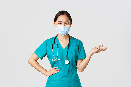 Covid-19, coronavirus disease, healthcare workers concept. Confused and upset asian female doctor in medical mask and scrubs, raise hand up puzzled, cant understand what problem, white background.