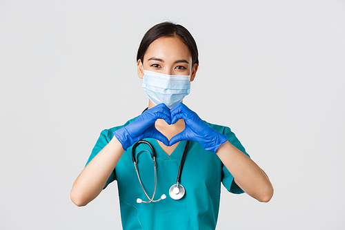 Covid-19, coronavirus disease, healthcare workers concept. Close-up of charming smiling asian female doctor, physician in medical mask and rubber gloves, provide care for patients, show heart gesture.
