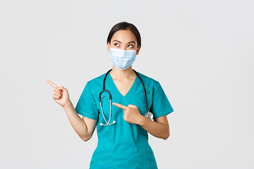 Covid-19, coronavirus disease, healthcare workers concept. Curious pleased asian female doctor, physician in medical mask and gloves, looking and pointing upper left corner with intrigued smile.