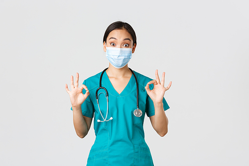 Covid-19, coronavirus disease, healthcare workers concept. Excited and impressed asian female doctor, intern in medical mask and scrubs showing okay gesture in approval, white background.