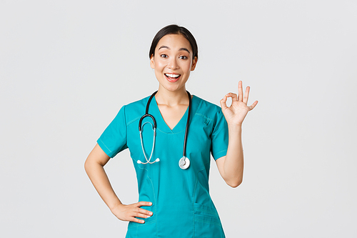 Covid-19, healthcare workers, pandemic concept. Surprised and happy asian female doctor, nurse in scrubs showing okay gesture and smiling amazed, praise nice work, agree with someone.
