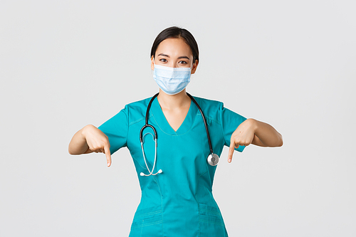 Covid-19, coronavirus disease, healthcare workers concept. Pleasant smiling asian female nurse, doctor in medical mask and scrubs, pointing fingers down, provide information, invite for checkup.