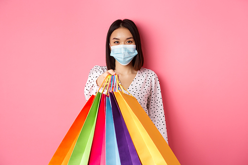 Covid-19, pandemic and lifestyle concept. Beautiful asian woman in face mask giving shopping bags, social distance in store, standing over pink background.