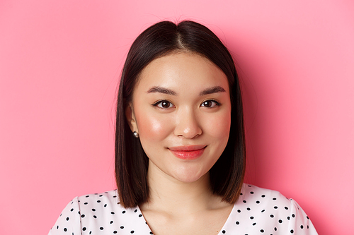 Beauty and lifestyle concept. Headshot of beautiful asian woman smiling, looking at camera happy and romantic, standing against pink background.