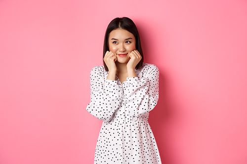 Cute and shy asian girl blushing, touching cheeks and looking left at copy space silly, standing against pink background.