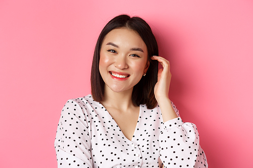 Close-up of beautiful asian woman smiling happy, touching new haircut, standing over pink background.