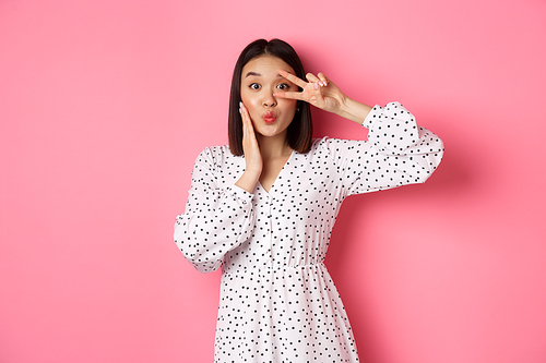 Coquettish asian woman in dress showing peace sign on eye, pucker lips for kiss, standing silly against pink background.