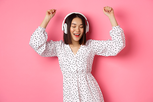 Happy young asian woman dancing and having fun, listening music in headphones, standing over pink background.