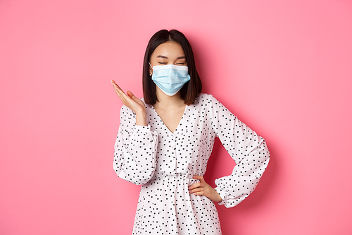 Covid-19, quarantine and lifestyle concept. Beautiful and confident asian woman in dress and face mask looking self-assured, standing sassy against pink background.