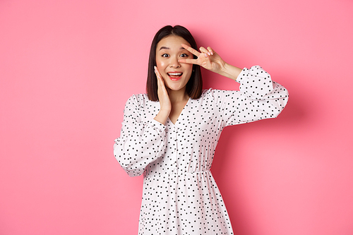 Beautiful and feminine asian woman showing kawaii sign on eye and looking amazed at camera, standing in dress over pink background.