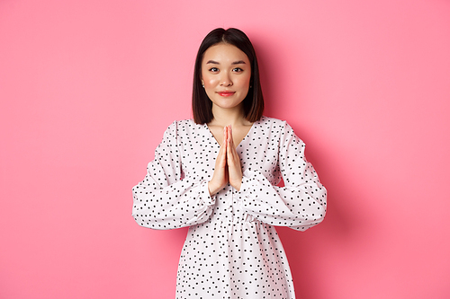 Beautiful asian lady in dress asking for help, holding hands in pray or namaste gesture, thanking you, standing over pink background.