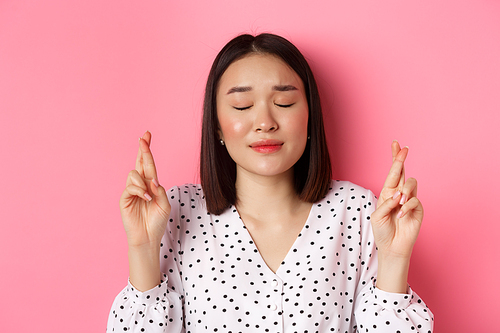 Beauty and lifestyle concept. Close-up of cute asian girl making a wish, close eyes and cross fingers hopeful, praying and standing over pink background.