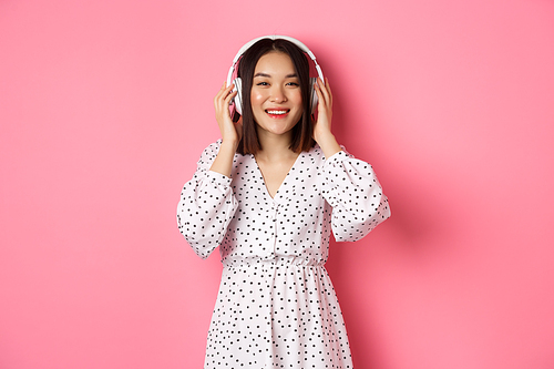 Romantic asian woman smiling happy, listening music in headphones and looking at camera, standing over pink background.