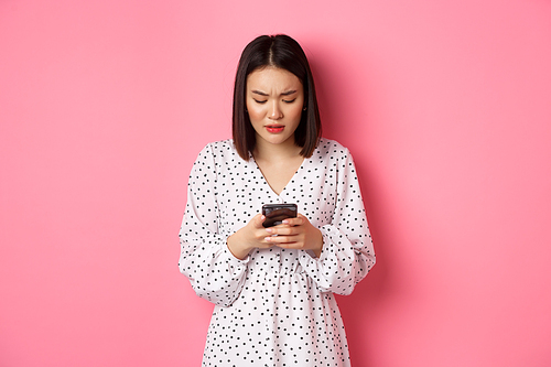 Nervous and concerned asian woman reading message on smartphone, looking worried, standing in dress over pink background.