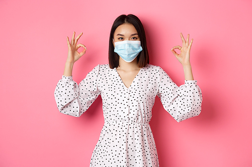 Coronavirus, social distancing and lifestyle concept. Satisfied asian woman in face mask showing okay signs, approve and agree, praising good choice, pink background.