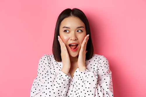 Close-up of beautiful asian woman looking surprised and excited, hear amazing news, looking left and rejoicing, standing against pink background.