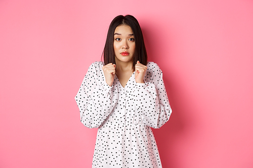 Scared cute asian girl in dress looking worried, feeling frightened and staring at camera nervously, standing over pink background.