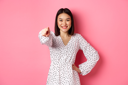 Confident asian woman in dress pointing at you, smiling and staring at camera, standing against pink background. Copy space