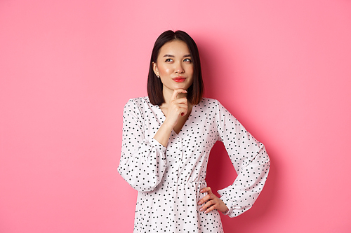 Thoughtful happy asian woman making decision, smiling satisfied and looking at upper left logo, thinking or choosing, standing in dress against pink background.