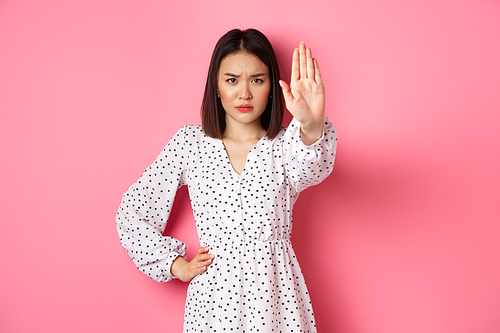 Angry asian woman tell stop, extend arm to prohibit or disapprove something, frowning displeased, standing over pink background.