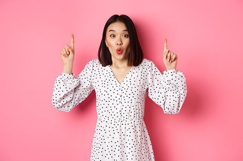 Pretty asian female model in dress pointing fingers up, showing copy space and staring at camera amazed, telling big news, standing over pink background.