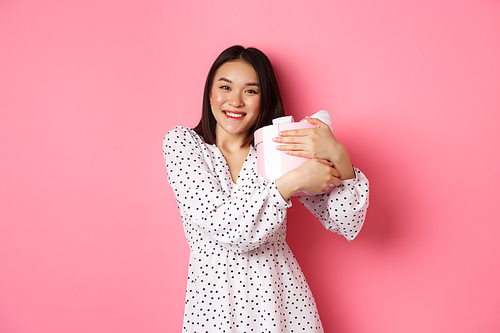 Lovely asian woman hugging her gift and smiling thankful, receive valentines day present, standing over pink background.