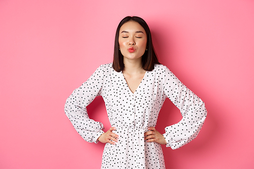 Beautiful asian woman in dress pucker lips and close eyes, waiting for kiss, standing over pink background.