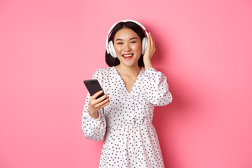 Beautiful asian woman listening music in headphones, using mobile phone, smiling happy at camera, standing over pink background.