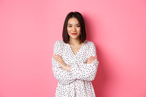 Beautiful asian female model standing in dress, cross arms on chest and smiling at camera, standing over pink background.