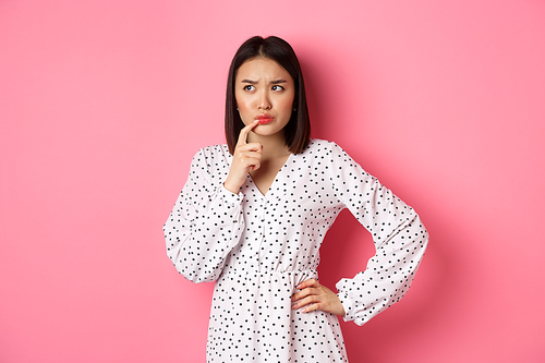 Troubled asian girl making decision, frowning and touching lip while thinking, looking unsure at upper left corner and choosing, standing over pink background.